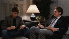 a man in a suit sits next to another man in a chair with a lamp in the background