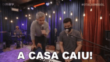 a man singing into a microphone next to another man playing a keyboard with the words a casa caiu written on the bottom