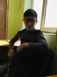 a boy wearing glasses is sitting at a desk in front of a window