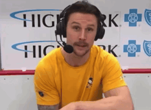 a man wearing headphones and a yellow shirt stands in front of a sign that says high