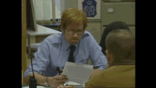 a man in a blue shirt and tie is sitting at a desk with a sticker on the wall that says police