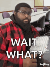 a man wearing glasses and a plaid shirt is holding a laptop and says wait what ?