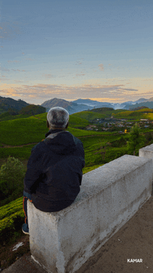 a man sits on a wall overlooking a valley and the word kamar is on the bottom right