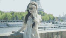 a woman wearing sunglasses and a white dress is standing on a bridge in front of the eiffel tower in paris .