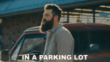 a man with a beard is standing in front of a red truck with the words " in a parking lot " below him