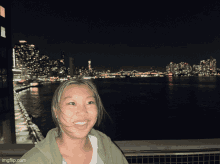 a woman stands in front of a body of water at night with a city skyline in the background