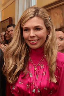 a woman wearing a pink dress and necklaces is smiling in front of a group of people .