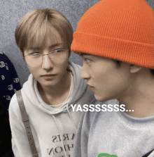 a man wearing glasses and an orange beanie looks at another man wearing a grey sweatshirt