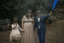 a man in a suit holds a blue smoke bomb while standing next to a pregnant woman and a little girl