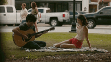 a man is playing a guitar to a woman while they are sitting on a blanket in a park .