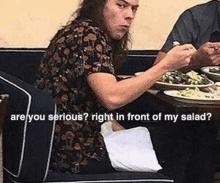 harry styles is sitting at a table eating a salad and talking to someone .