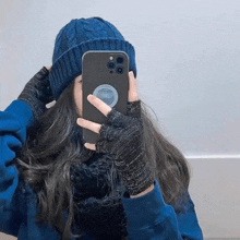 a woman wearing a blue hat and gloves is taking a selfie with her cell phone .