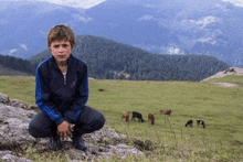 a young boy in a blue adidas jacket squatting on a rock