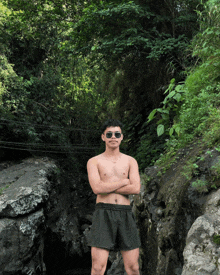 a shirtless man in shorts stands with his arms crossed in front of a forest