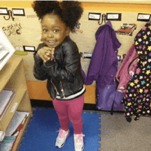 a little girl wearing a leather jacket and pink pants stands in front of a wall that says speed