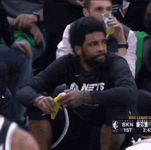 a basketball player wearing a nets shirt sits in the stands eating a banana