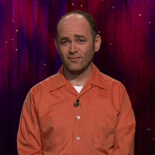 a man in an orange shirt with a microphone on his neck stands in front of a red curtain