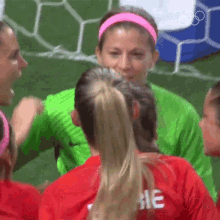 a group of female soccer players are huddled together on the field . one of the players is wearing a pink headband .