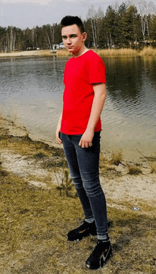 a young man in a red t-shirt is standing in front of a body of water