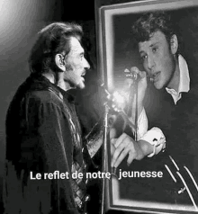 a black and white photo of a man singing into a microphone and looking at his reflection .