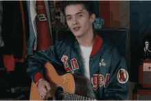 a young man is playing a guitar and singing while wearing a jacket with the word angels on it .