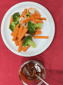 a plate of carrots broccoli and shrimp next to a bowl of barbecue sauce