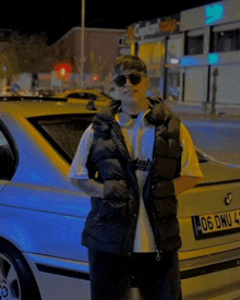 a man wearing sunglasses and a black vest is standing in front of a car with a license plate that says 06 onu 4