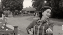 a black and white photo of a woman in a plaid dress standing on a sidewalk in front of a house .