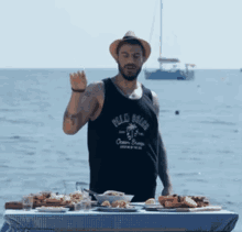 a man wearing a black tank top that says " belle beach " stands in front of a table full of food