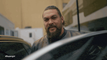 a man with a beard is standing next to a car with the hashtag #supergirl on the bottom