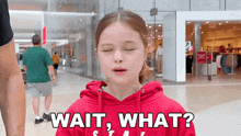 a little girl wearing a red hoodie says wait , what ? in a mall .