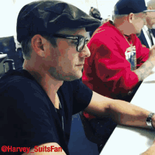 a man wearing glasses and a flat cap sits at a table with a picture of him taken by harvey suitsfan