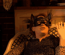 a woman wearing a cat mask sits in a chair with her hand up