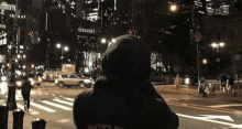 a person in a hoodie is standing on a city street at night looking at traffic .