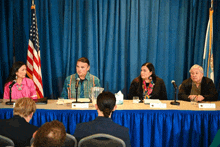 a group of people sit at a table with microphones in front of a blue curtain