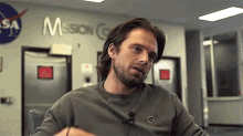 a man with long hair and a beard is sitting at a table in front of a nasa sign .