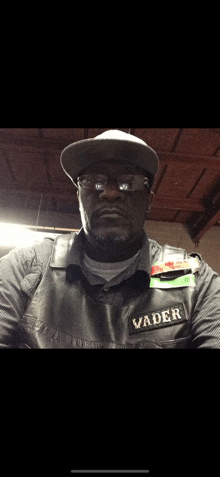 a man wearing a hat and glasses has the word vader on his vest