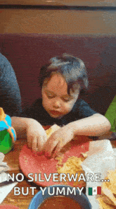 a young boy is sitting at a table with a plate of food and a sign that says no silverware but yummy .