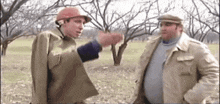 two men are standing in a field and talking to each other . one of the men is wearing a hard hat .