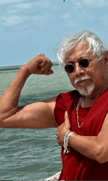 a man wearing sunglasses and a red tank top is flexing his muscles in front of the ocean .