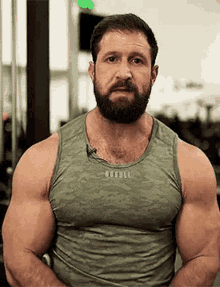 a man with a beard and a green tank top is standing in a gym with his arms crossed .