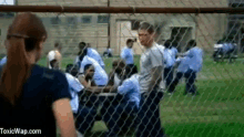 a woman is standing in front of a chain link fence looking at a group of prisoners behind it .