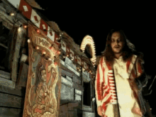 a man with long hair and a beard is standing in front of a carnival tent holding a cane and a sign .