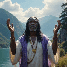 a man with long hair and a beard is standing in front of a body of water with his hands in the air