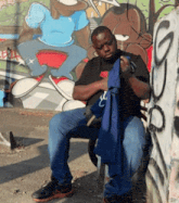 a man is sitting in front of a wall with graffiti on it and holding a blue shirt that says ' nc '