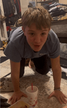 a boy in a blue shirt is crawling on the floor next to a cup with a red straw