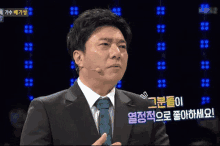 a man in a suit and tie speaking into a microphone with korean writing on the screen behind him