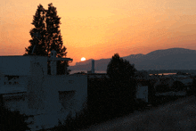 a sunset with a building in the foreground and the sun shining through the trees