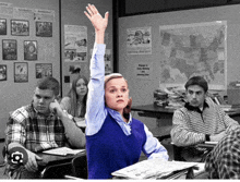 a girl in a blue vest raises her hand in a classroom with other students