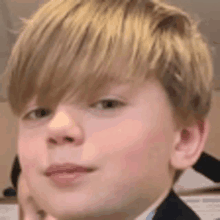 a close up of a young boy 's face with blonde hair and a black suit .